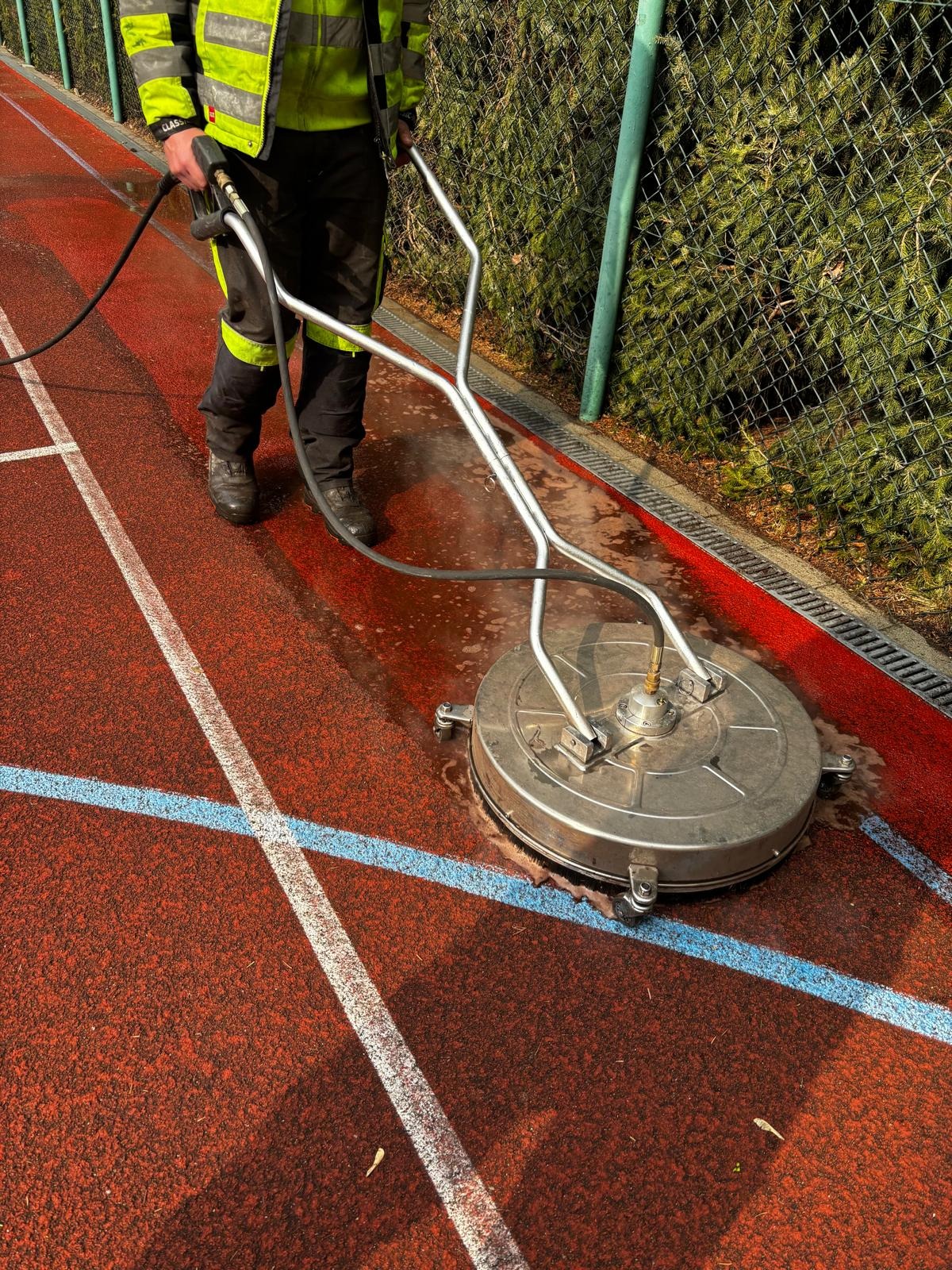 Czyszczenie czerwonej nawierzchni tartanowej boiska sportowego za pomocą okrągłej myjki ciśnieniowej z metalową obudową, obsługiwanej przez pracownika w odblaskowej kamizelce, na tle zielonego...