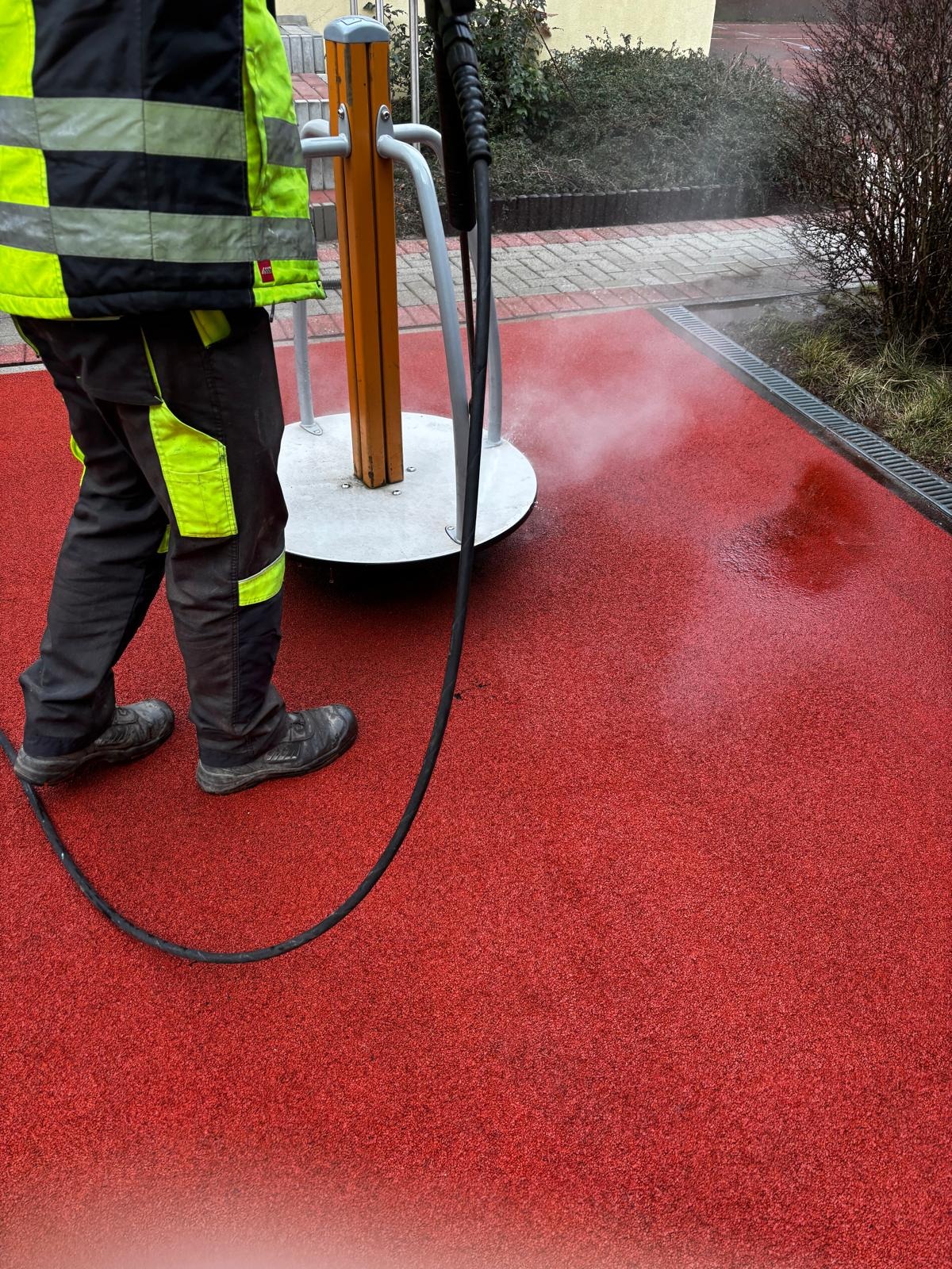 Pracownik w odblaskowej odzieży roboczej czyści czerwoną, gumowaną nawierzchnię placu zabaw za pomocą myjki ciśnieniowej, obok widoczny element zabawowy.