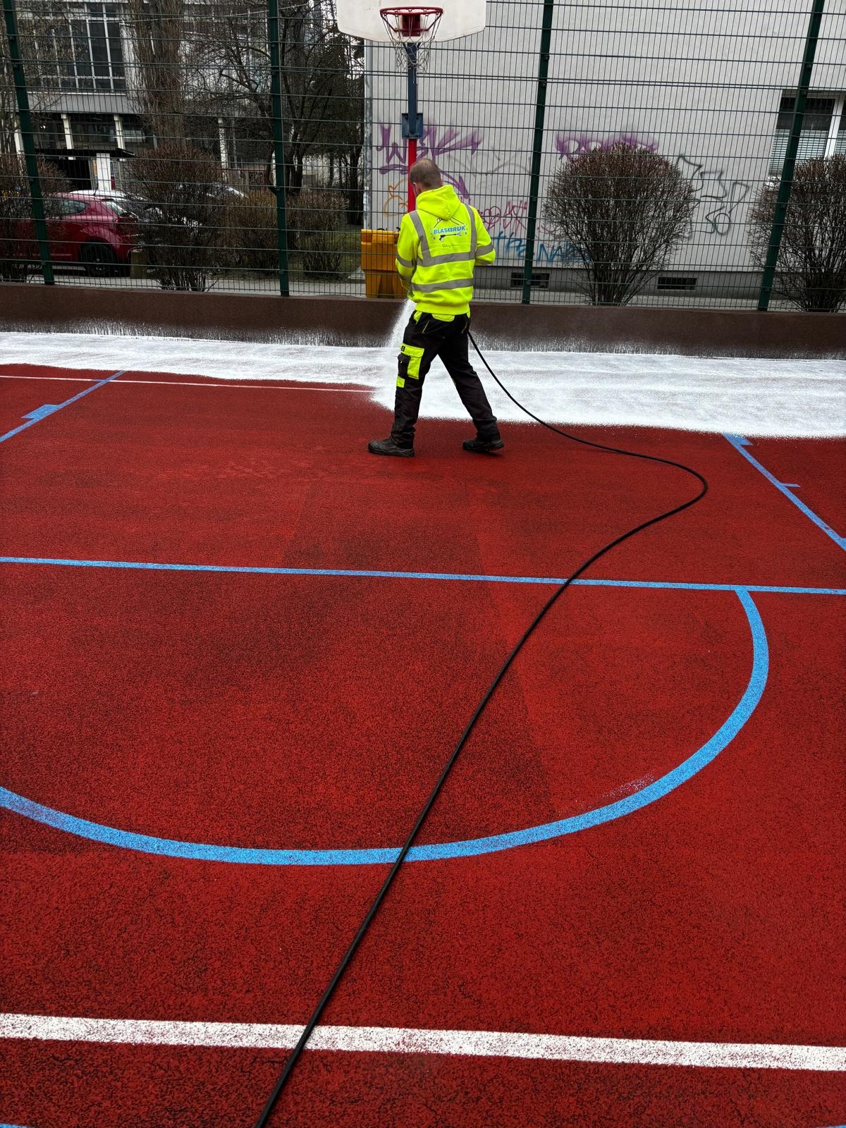 Pracownik w żółtej kamizelce roboczej czyści czerwoną nawierzchnię boiska sportowego z niebieskimi i białymi liniami za pomocą myjki ciśnieniowej, w tle widoczny budynek i ogrodzenie.