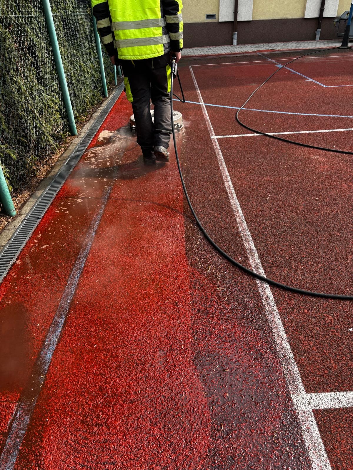 Pracownik w odblaskowej kamizelce czyści czerwoną nawierzchnię boiska sportowego za pomocą rotacyjnej maszyny czyszczącej, widoczne ślady po czyszczeniu i odpływ wody do kanalizacji liniowej.