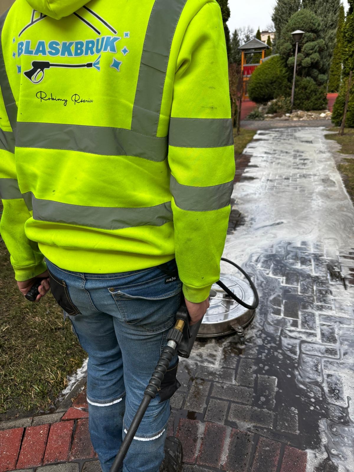 Pracownik w żółtej bluzie z logo firmy Blaskbruk czyści kostkę brukową za pomocą myjki ciśnieniowej z okrągłą dyszą, w tle widoczny plac zabaw.