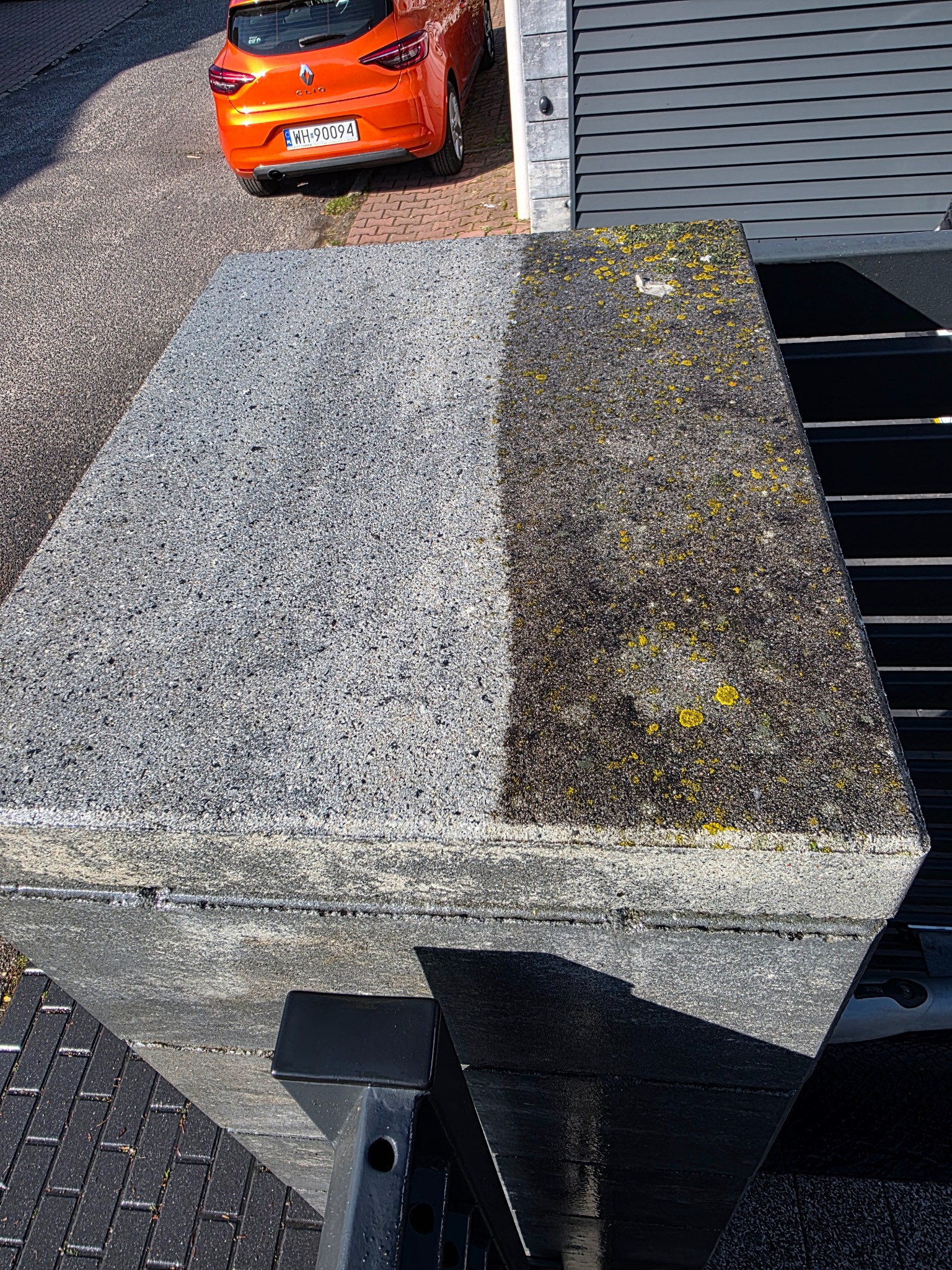 Fragment betonowej kostki brukowej, połowa wyczyszczona z mchu i zabrudzeń, widoczna różnica przed i po zabiegu, pomarańczowy samochód w tle.