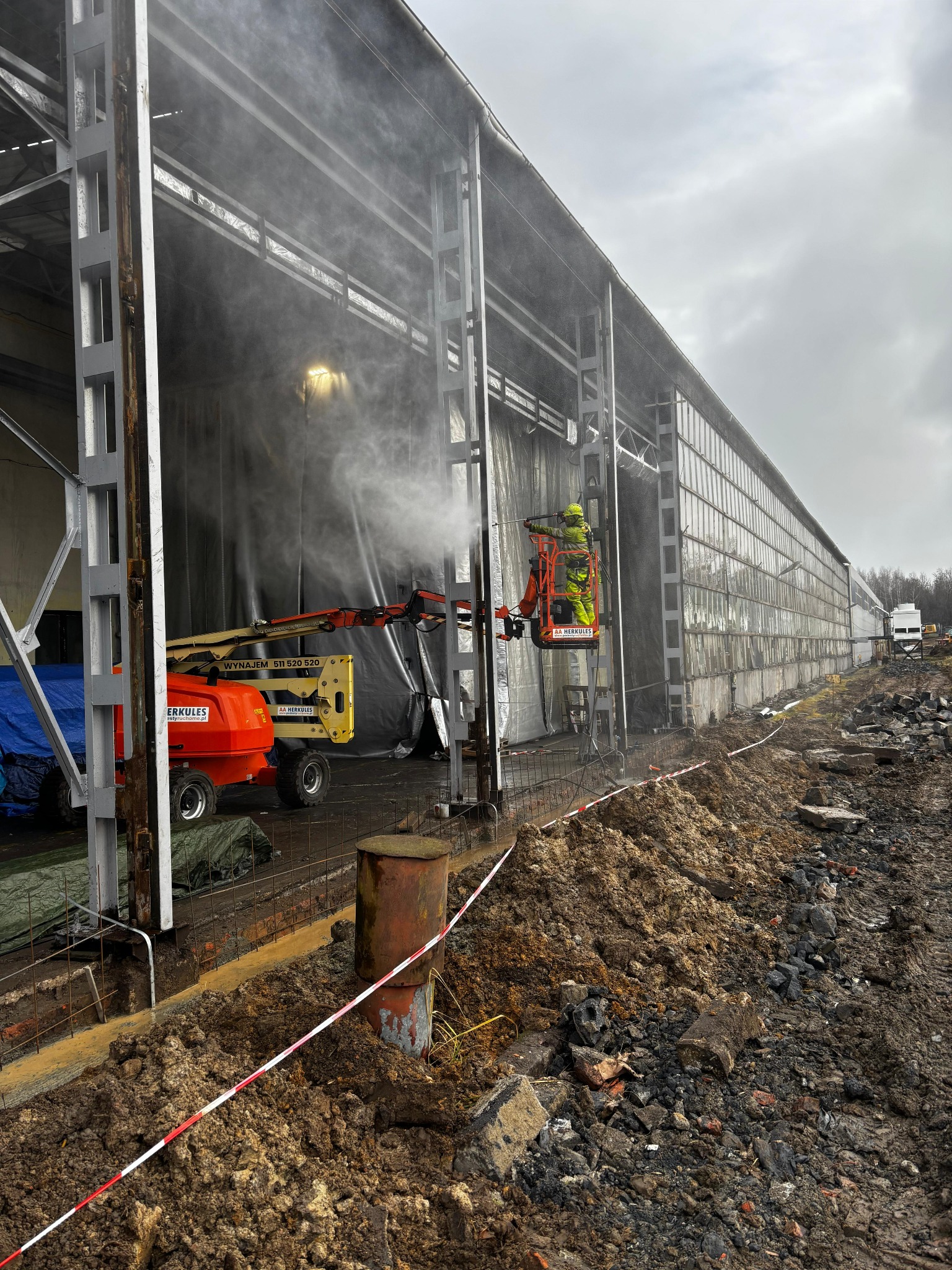 Pracownik w podnośniku koszowym czyści elewację hali przemysłowej strumieniem wody pod ciśnieniem. Widoczny sprzęt Herkules i teren budowy.