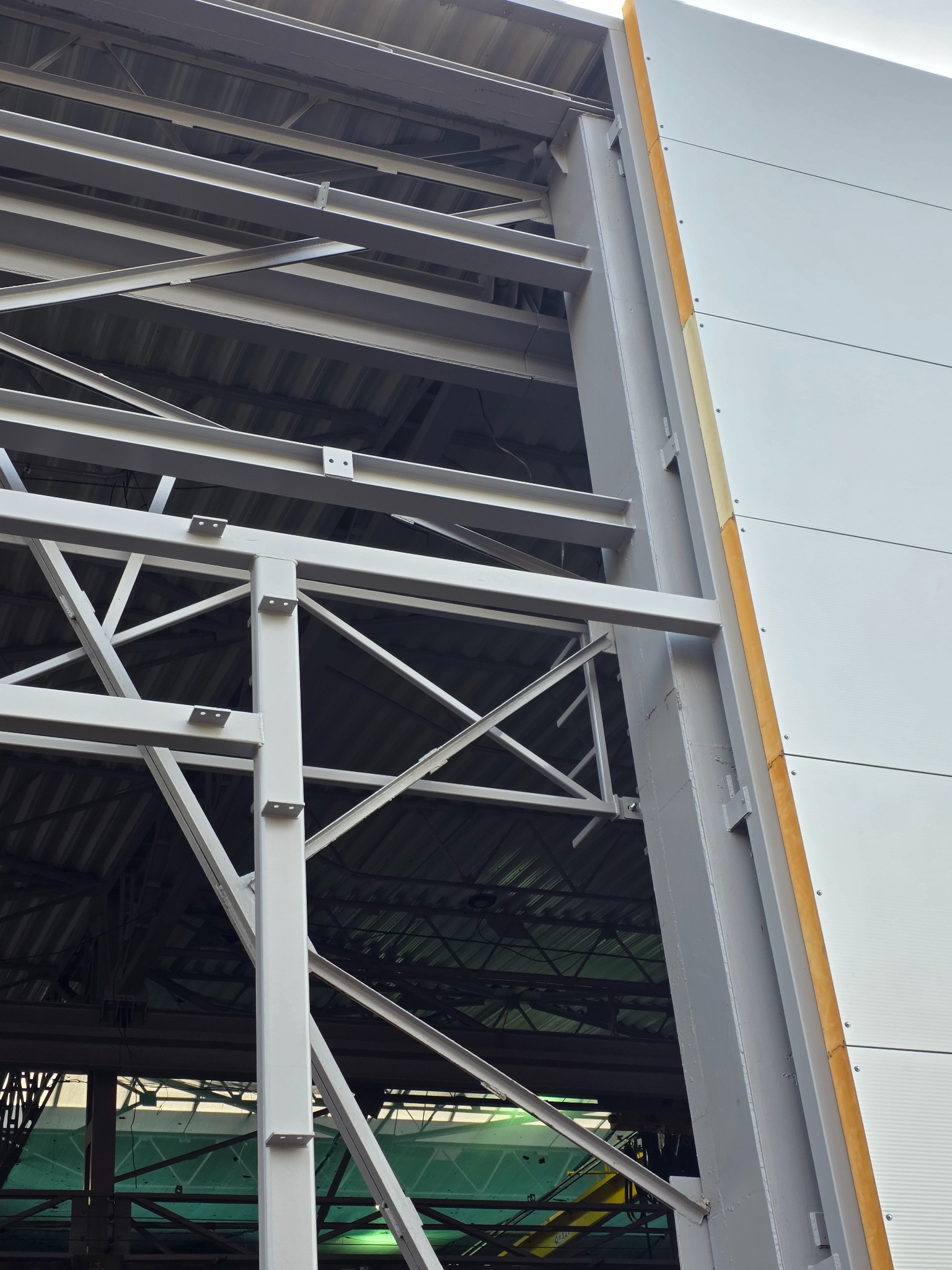 Szara stalowa konstrukcja hali przemysłowej z widocznymi elementami dachu i fragmentem elewacji z paneli. Widok z dołu, podkreślający solidność i precyzję wykonania.