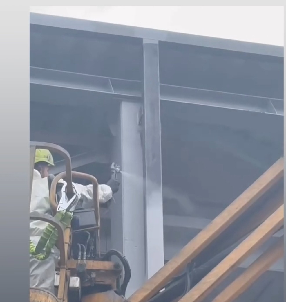 Pracownik w kombinezonie maluje konstrukcję stalową natryskowo z podnośnika koszowego w Mstowie. Widoczne rozpylanie farby i elementy konstrukcji budynku.