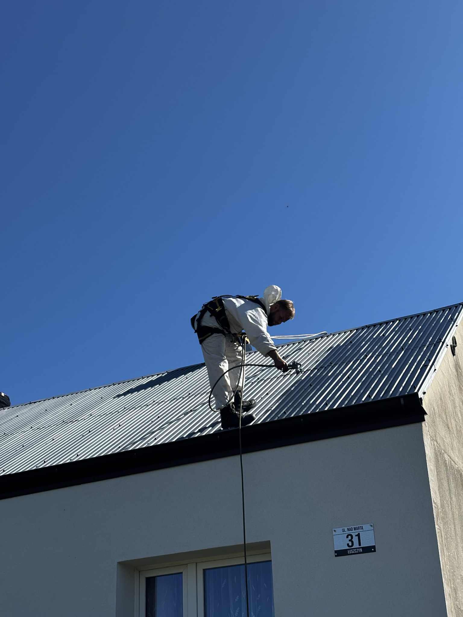 Pracownik w kombinezonie maluje metalowy dach natryskowo, zabezpieczony linami. Widoczny adres budynku: ul. Nad Wartą 31, Łuszczyn. Błękitne niebo w tle.