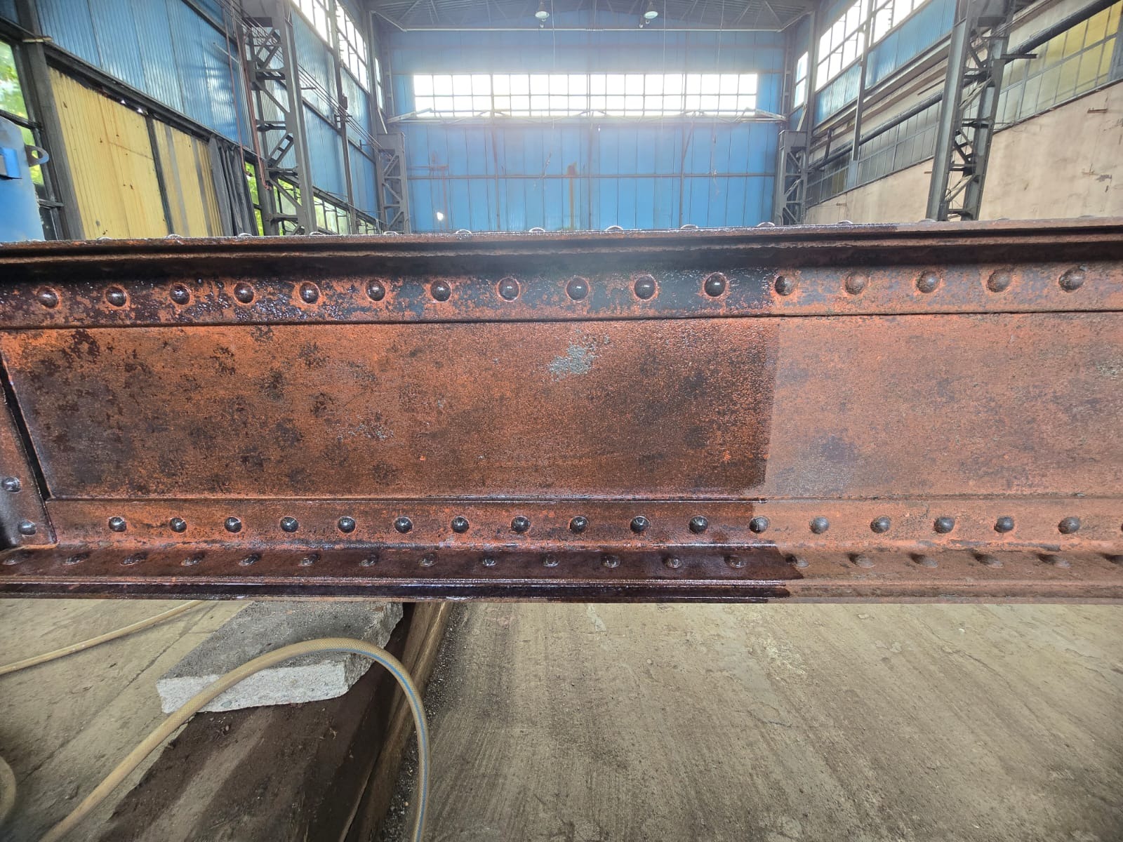 Metalowa konstrukcja w hali przemysłowej, częściowo oczyszczona z rdzy. Widoczna różnica między stanem przed i po czyszczeniu, z detalami nitów i fakturą metalu.