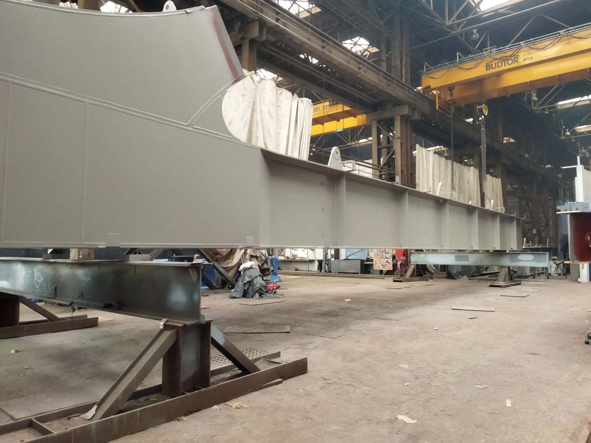 Szara, pomalowana konstrukcja stalowa na podporach w hali przemysłowej z suwnicą BUDTOR w tle. Widoczne elementy konstrukcyjne i oświetlenie hali.