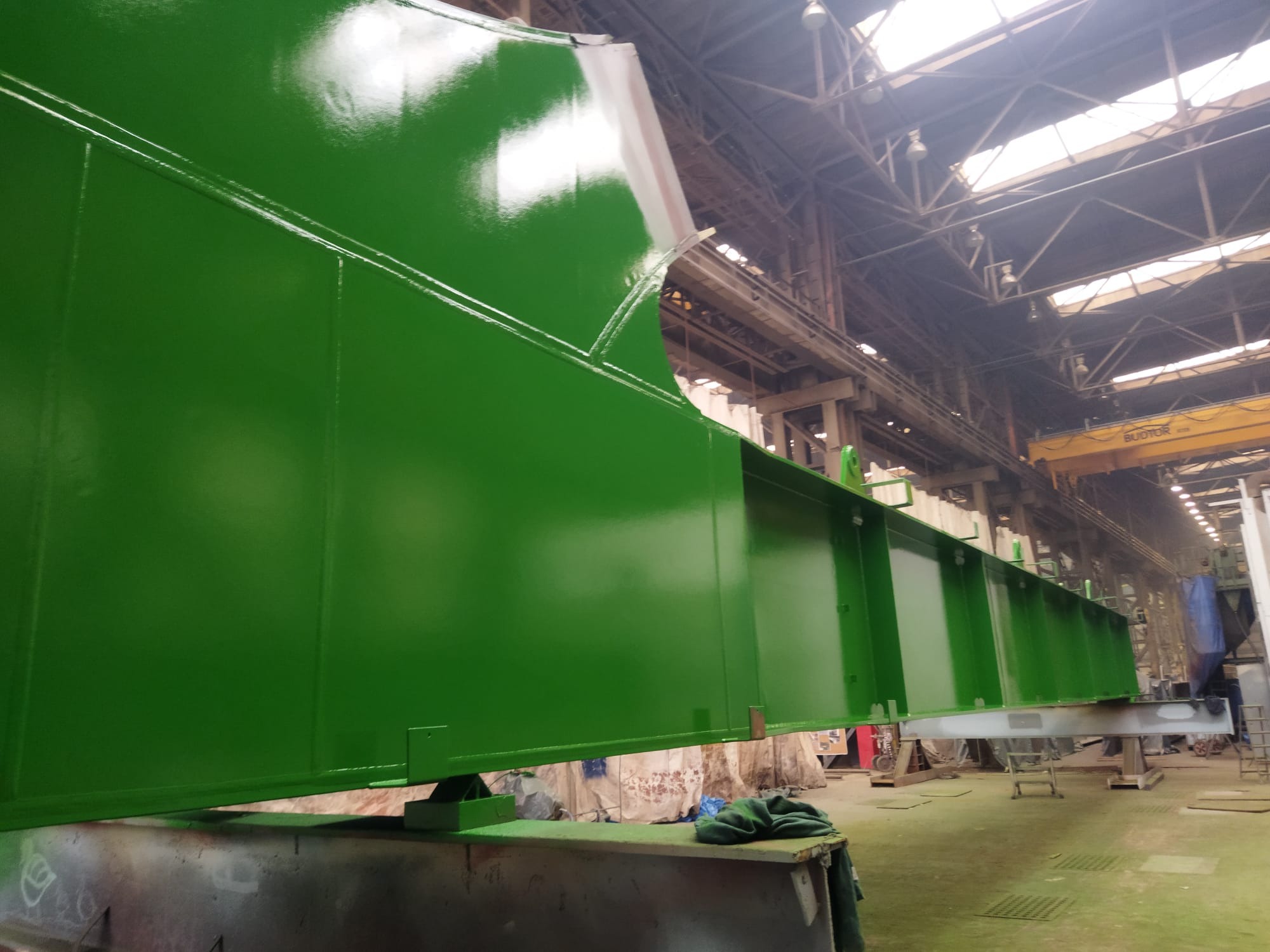 Pomarańczowo-zielona, świeżo pomalowana konstrukcja stalowa w hali przemysłowej, widok z dołu, oświetlenie górne, suwnica w tle.