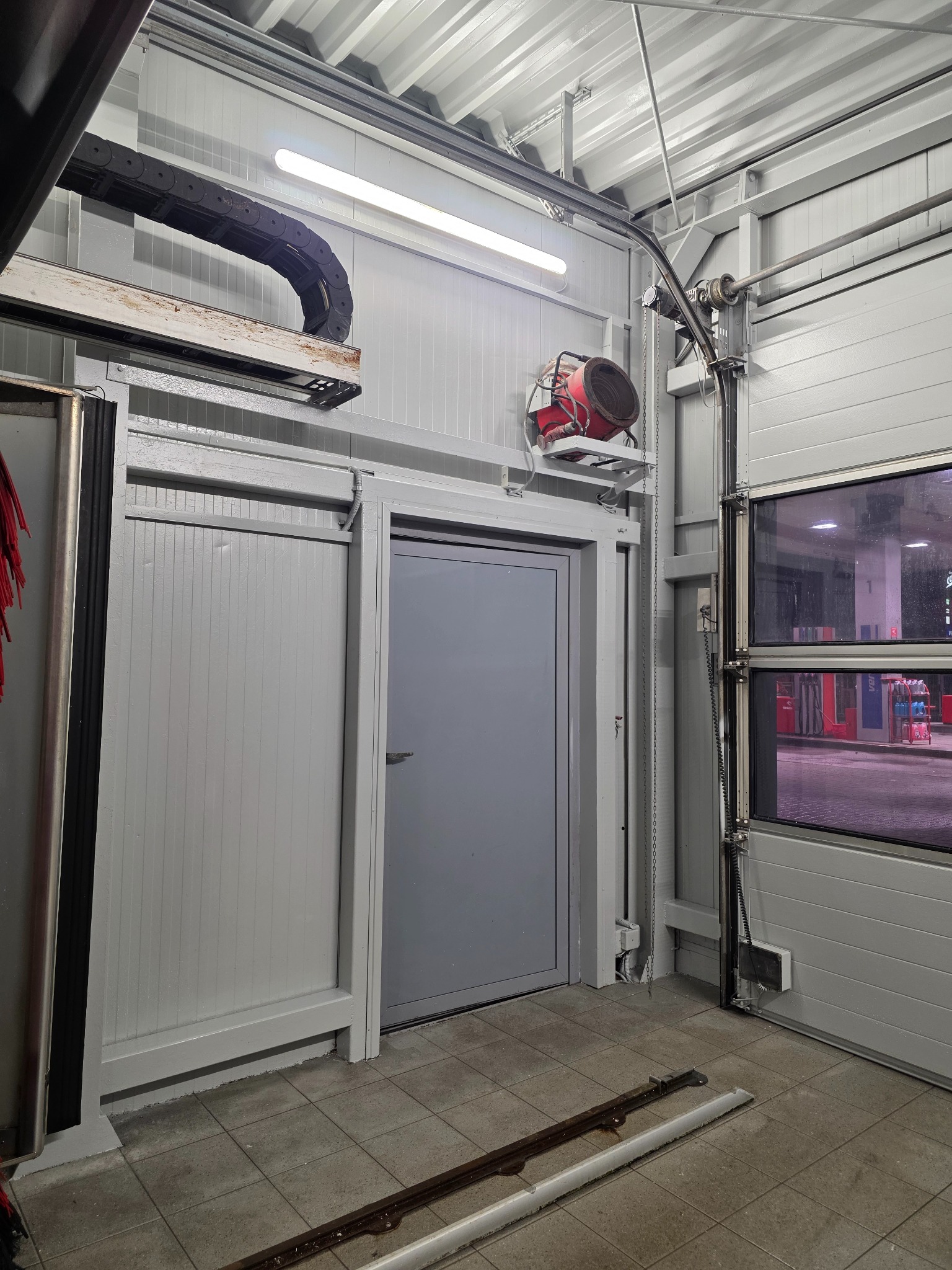 Wnętrze hali z szarą bramą segmentową i drzwiami. Widoczne elementy konstrukcyjne, oświetlenie i fragment myjni samochodowej. W tle stacja benzynowa.