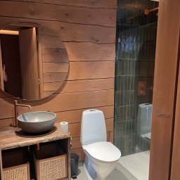 Stolarstwo - Drewniana łazienka z okrągłym lustrem, kamienną umywalką i rattanowymi koszami. Kabina prysznicowa z zielonymi płytkami w tle. Ciepłe oświetlenie.