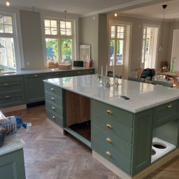 Stolarstwo - Wnętrze kuchni w stylu klasycznym z zielonymi szafkami i marmurowym blatem wyspy. Widoczny parkiet w jodełkę i duże okna w tle. Częściowy montaż.