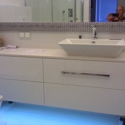 Stolarstwo - Biała szafka łazienkowa pod umywalką z podświetleniem LED, lustro z dekoracyjną ramą. Widoczne akcesoria łazienkowe i odbicie osoby robiącej zdjęcie.