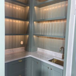 Stolarstwo - Szara zabudowa kuchenna z marmurowym blatem, złotą armaturą i oświetleniem LED w półkach. Klasyczny styl, widok z perspektywy wejścia do pomieszczenia.