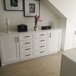 Stolarstwo - Biała komoda z szufladami i szafkami, czarne uchwyty, dekoracje na blacie, drewniana podłoga, skos dachu, minimalistyczny styl, czarno-białe zdjęcia w ramkach na ścianie.