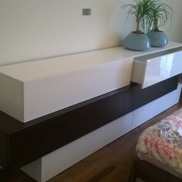 Stolarstwo - Nowoczesna szafka RTV w kolorach bieli i ciemnego brązu, z wysuwaną szufladą i dekoracyjnymi roślinami w niebieskich doniczkach na blacie, obok łóżko.