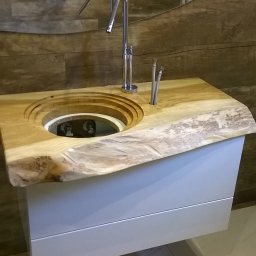 Stolarstwo - Unikatowa umywalka z blatem z litego drewna o nieregularnym kształcie, osadzona na białej szafce z szufladami. W tle brązowa ściana imitująca drewno i grzejnik.