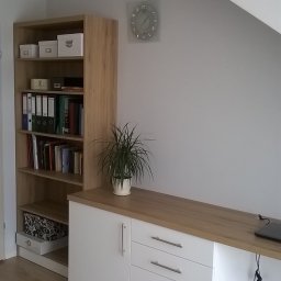 Stolarstwo - Białe szafki z drewnianym blatem i regał z książkami w pokoju na poddaszu. Na blacie doniczka z rośliną i laptop. Na ścianie zegar. Widoczne gniazdka elektryczne.