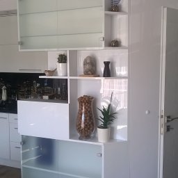 Stolarstwo - Biała, nowoczesna szafa wnękowa z półkami i przeszklonymi drzwiczkami, dekorowana elementami wystroju wnętrz. Widoczny fragment kuchni w tle.
