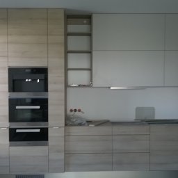 Stolarstwo - Jasna kuchnia z drewnianymi frontami i wbudowanym sprzętem AGD, minimalistyczny design, oświetlenie podszafkowe, widok na okno z lewej strony.