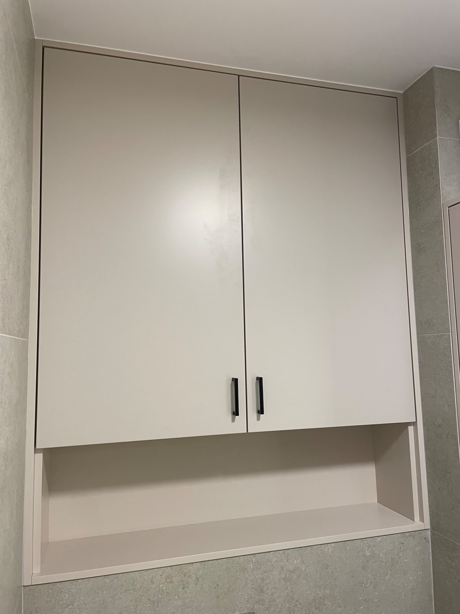 Zamknięta szafka łazienkowa w odcieniach beżu z otwartą półką na dole, minimalistyczne czarne uchwyty, na tle szarej ściany z płytek.