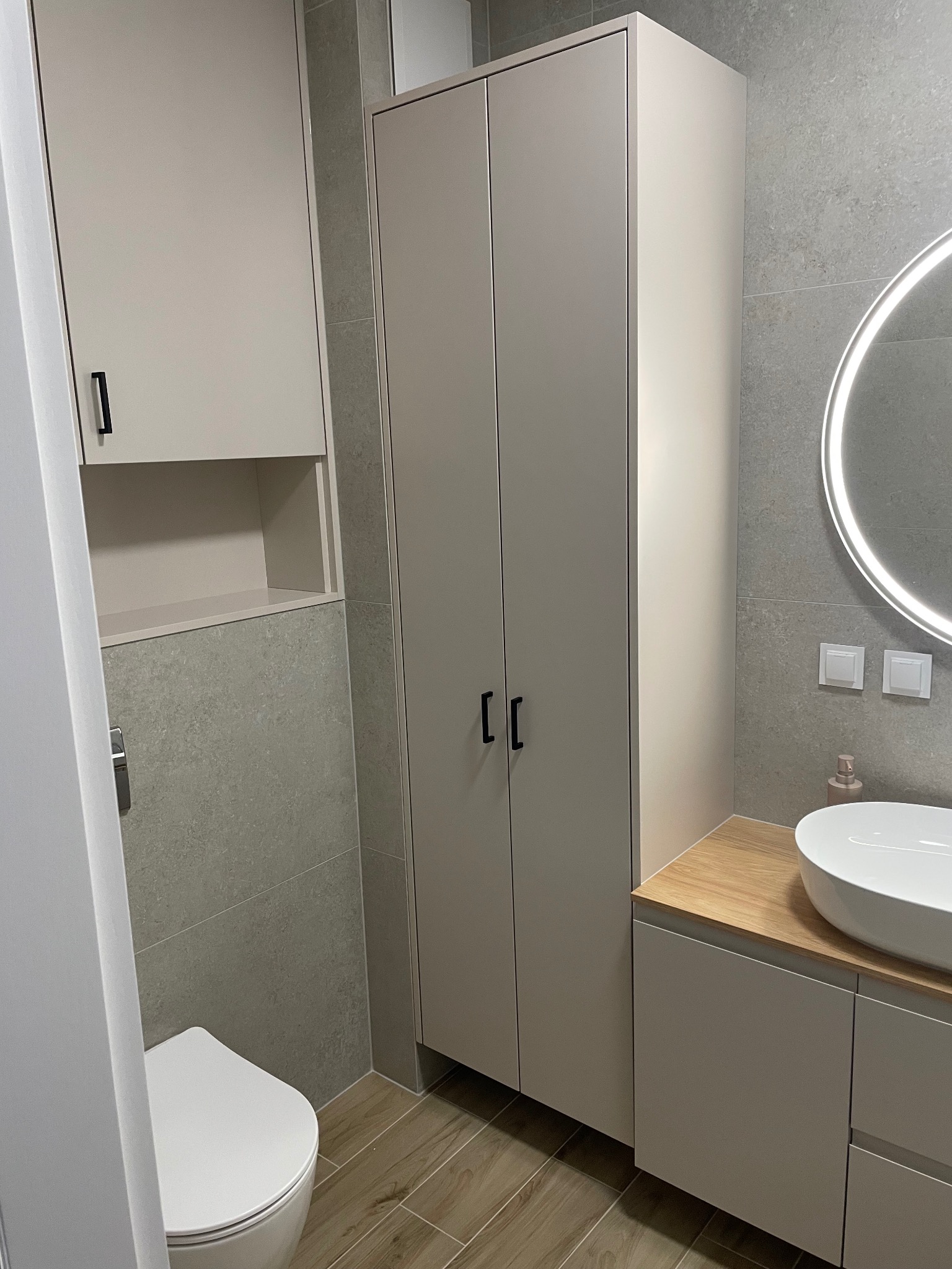 Nowoczesna łazienka z beżowymi szafkami, drewnianym blatem pod umywalką i okrągłym lustrem z podświetleniem LED. Minimalistyczny design, szare płytki na ścianach i podłodze.