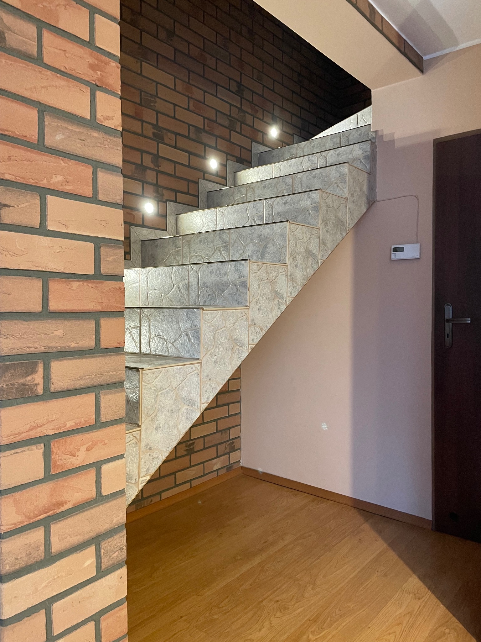 Schody z płytek z oświetleniem punktowym, obłożone cegłą w dwóch odcieniach, prowadzące do ciemnych drzwi. Parkiet na podłodze.