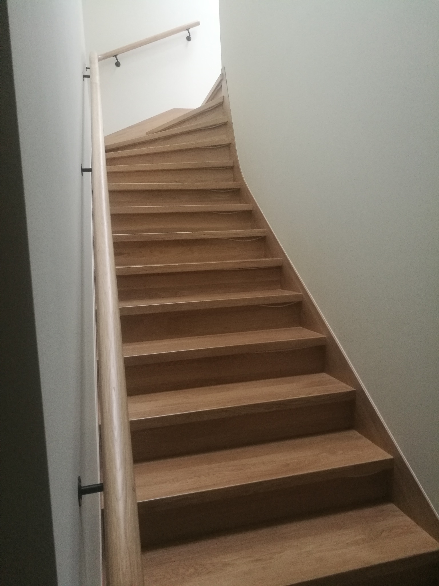 Drewniane schody z jasną poręczą zamontowaną do ściany, widok z dołu. Realizacja z Wejherowa. Stopnie schodów w naturalnym odcieniu drewna.