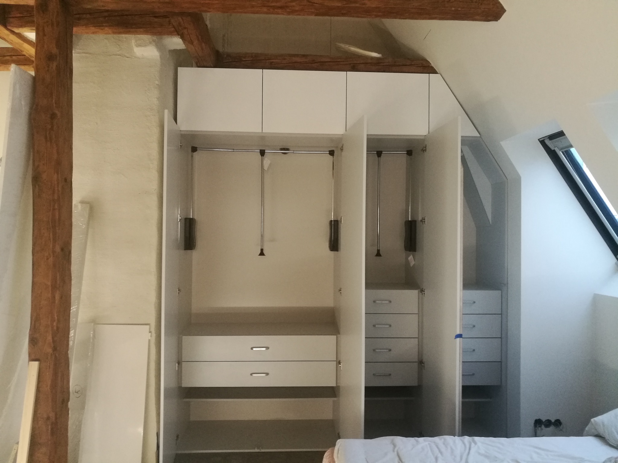 Biała szafa wnękowa z otwartymi drzwiami, widoczne półki, szuflady i drążki na ubrania, umieszczona na poddaszu ze skośnym oknem dachowym i drewnianą belką stropową.
