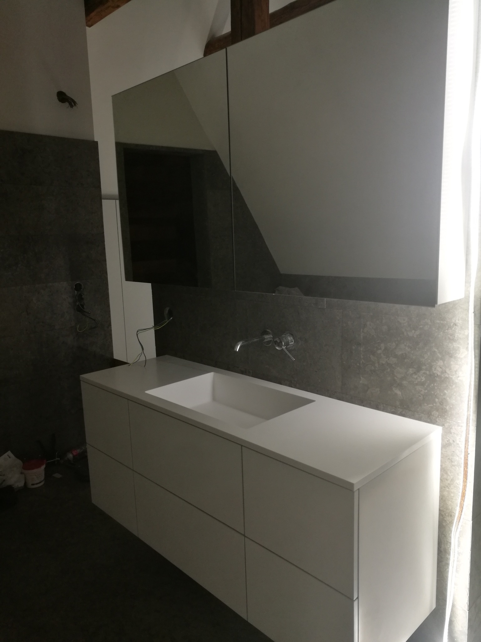 Nowoczesna, biała szafka łazienkowa z umywalką i lustrem na tle szarej ściany. Widoczne elementy instalacji elektrycznej. Minimalistyczny design.