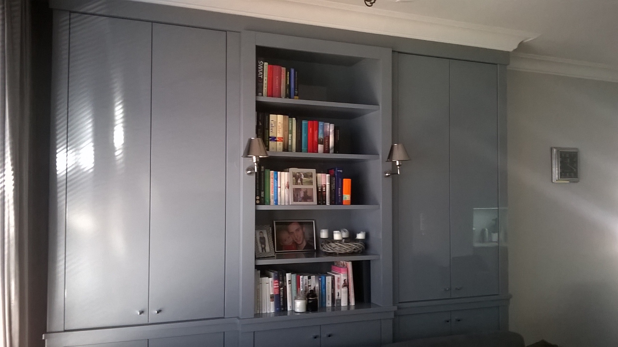 Szara zabudowa meblowa z szafkami i otwartą półką na książki, oświetlona kinkietami. Widoczne dekoracje i książki na półkach. Minimalistyczny design.