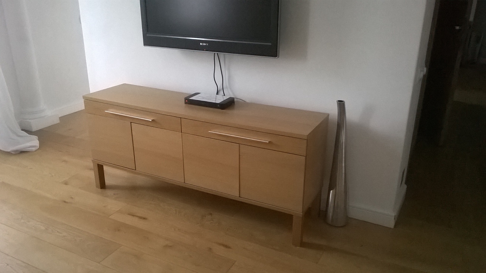 Drewniana komoda z szufladami i szafkami, z metalowymi uchwytami, pod telewizorem. Na blacie dekoder. Obok wazon. Parkiet na podłodze. Minimalistyczny styl.