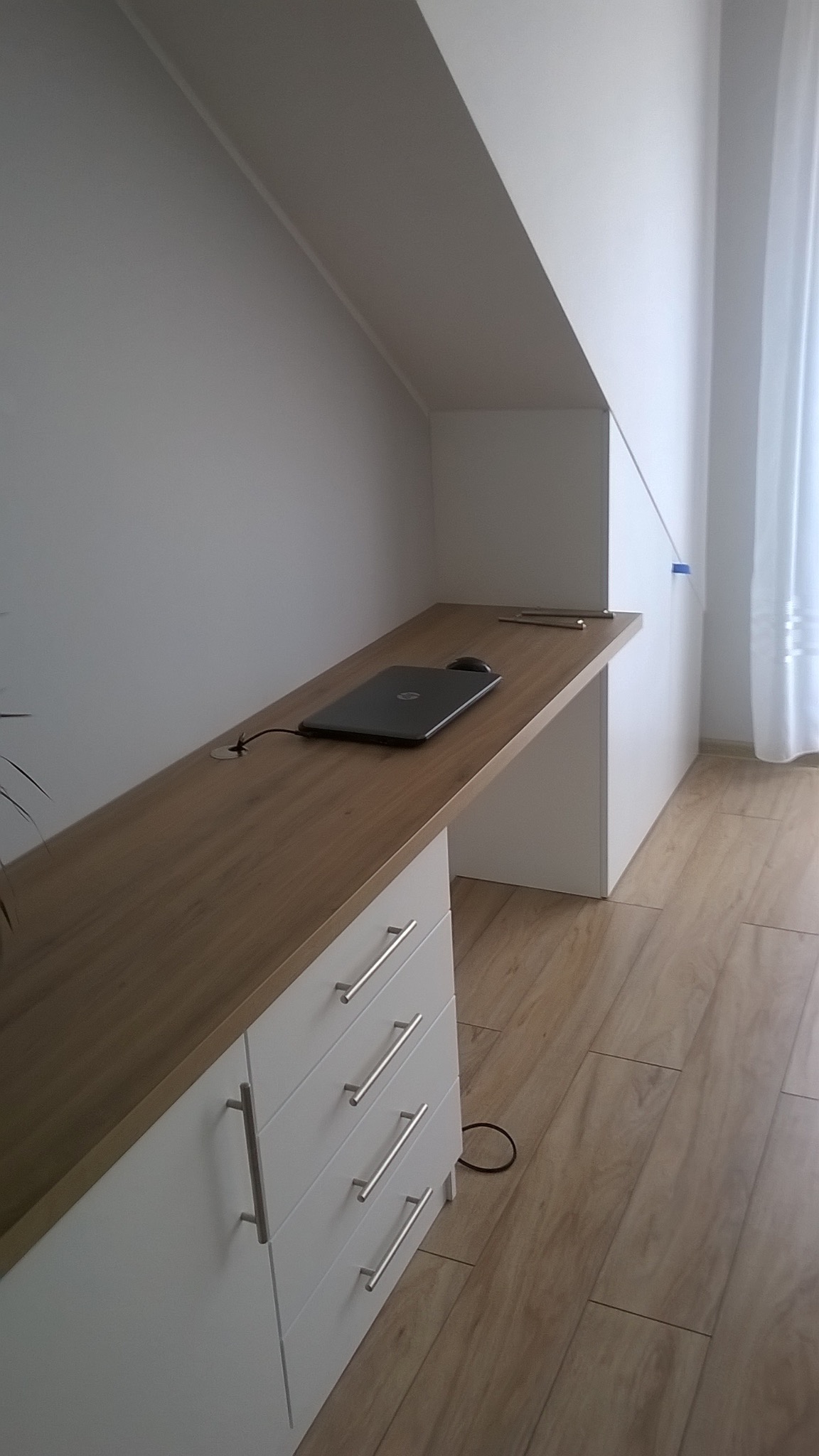 Drewniany blat biurka na szafce z szufladami, wkomponowany we wnękę pod skosem. Na blacie laptop i myszka. Jasne wnętrze, drewniana podłoga.