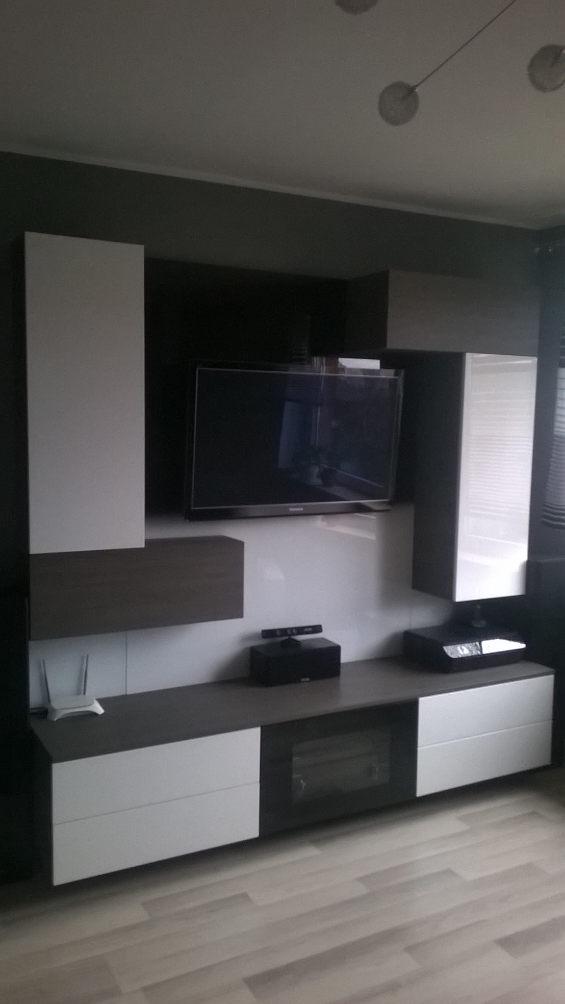 Nowoczesna zabudowa meblowa RTV w salonie, z białymi i grafitowymi frontami, wiszącymi szafkami i półkami. Na blacie sprzęt RTV. Minimalistyczny design.