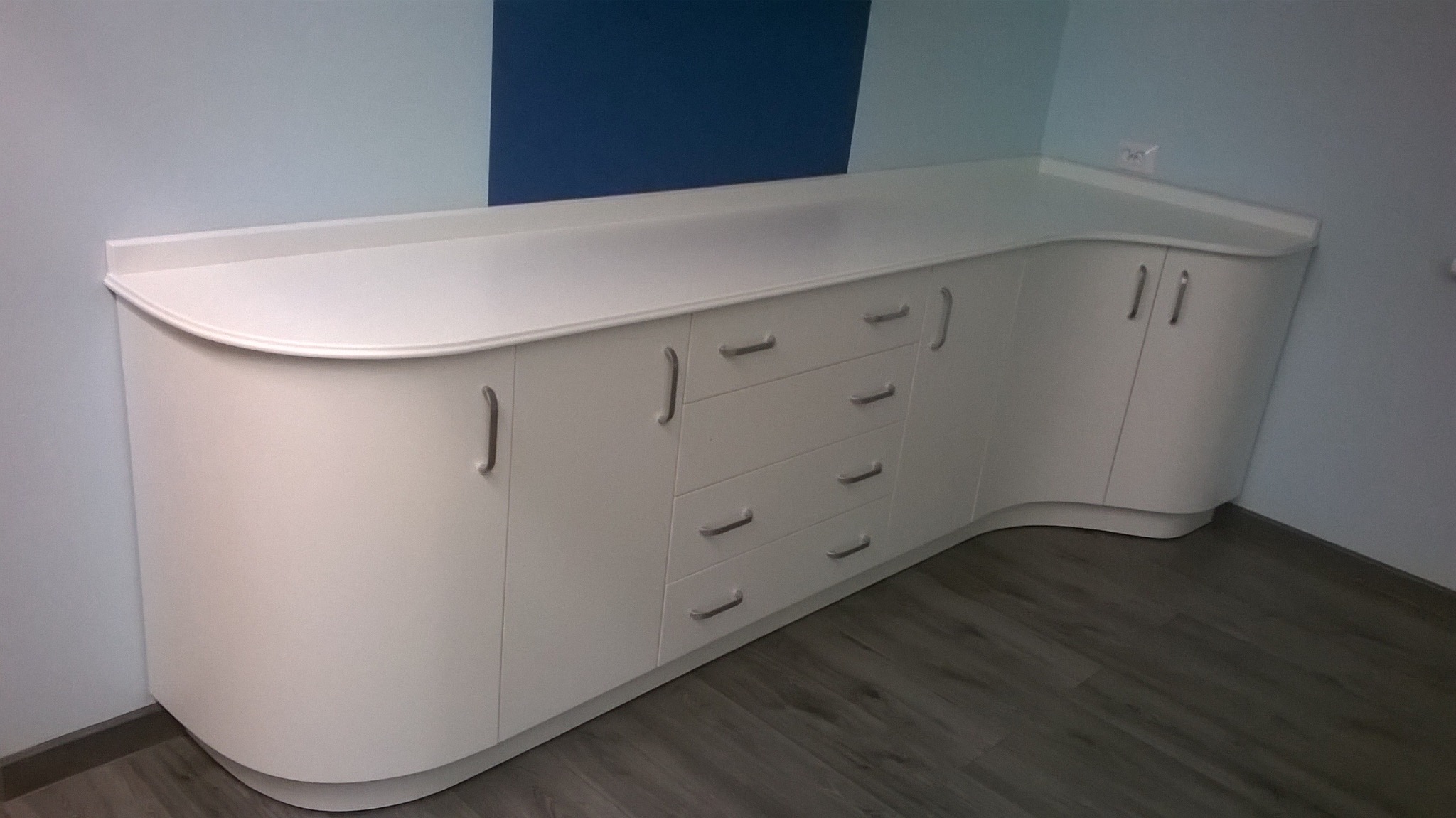 Biała, zaokrąglona szafka z szufladami i szafkami, z metalowymi uchwytami, na tle jasnoniebieskiej ściany z ciemnoniebieskim akcentem. Widoczna podłoga imitująca drewno.