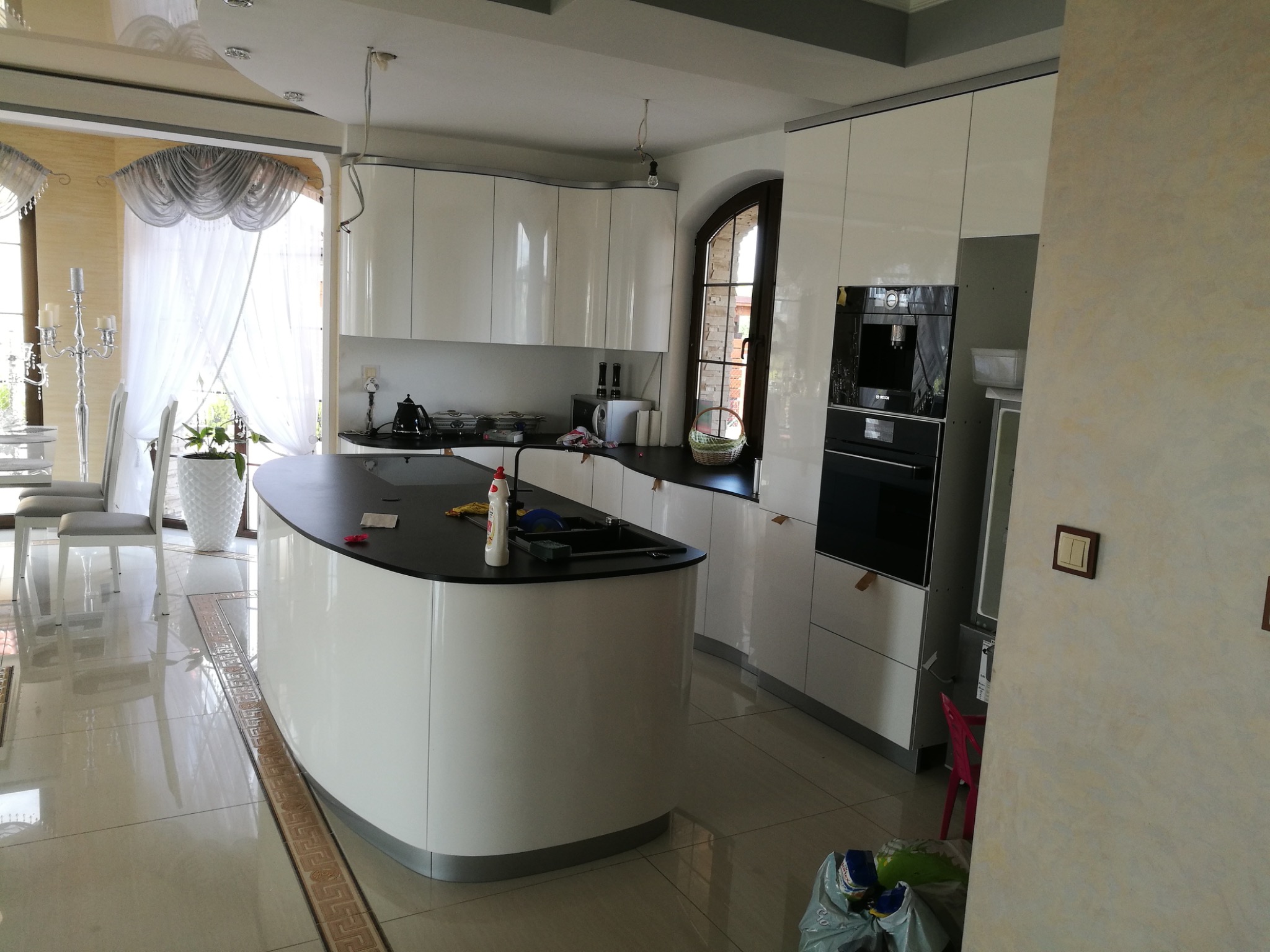 Nowoczesna kuchnia na wymiar z wyspą i białymi, lakierowanymi frontami. Widoczne wbudowane sprzęty AGD i okno łukowe. Błyszcząca podłoga z dekoracyjnym wzorem.