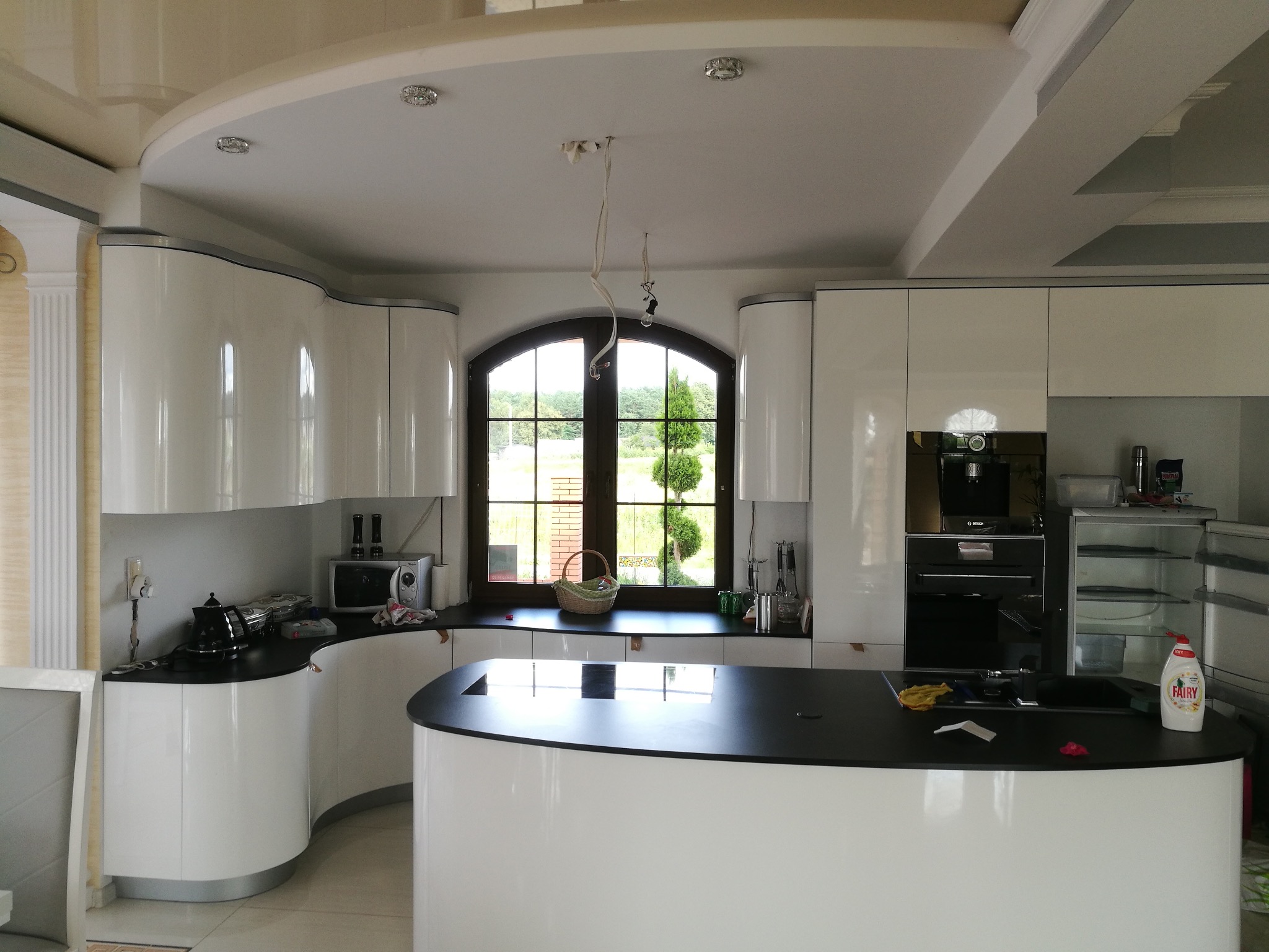 Nowoczesna kuchnia z białymi, zaokrąglonymi szafkami i czarnym blatem. Okno z widokiem na zieleń. Wiszące kable od lampy.