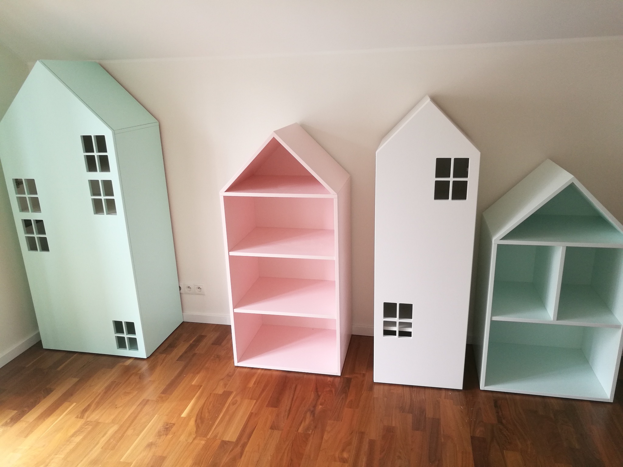 Kolorowe regały w kształcie domków, pastelowa zieleń, róż i biel, na drewnianej podłodze. Nietypowe meble do pokoju dziecięcego, oryginalny design.