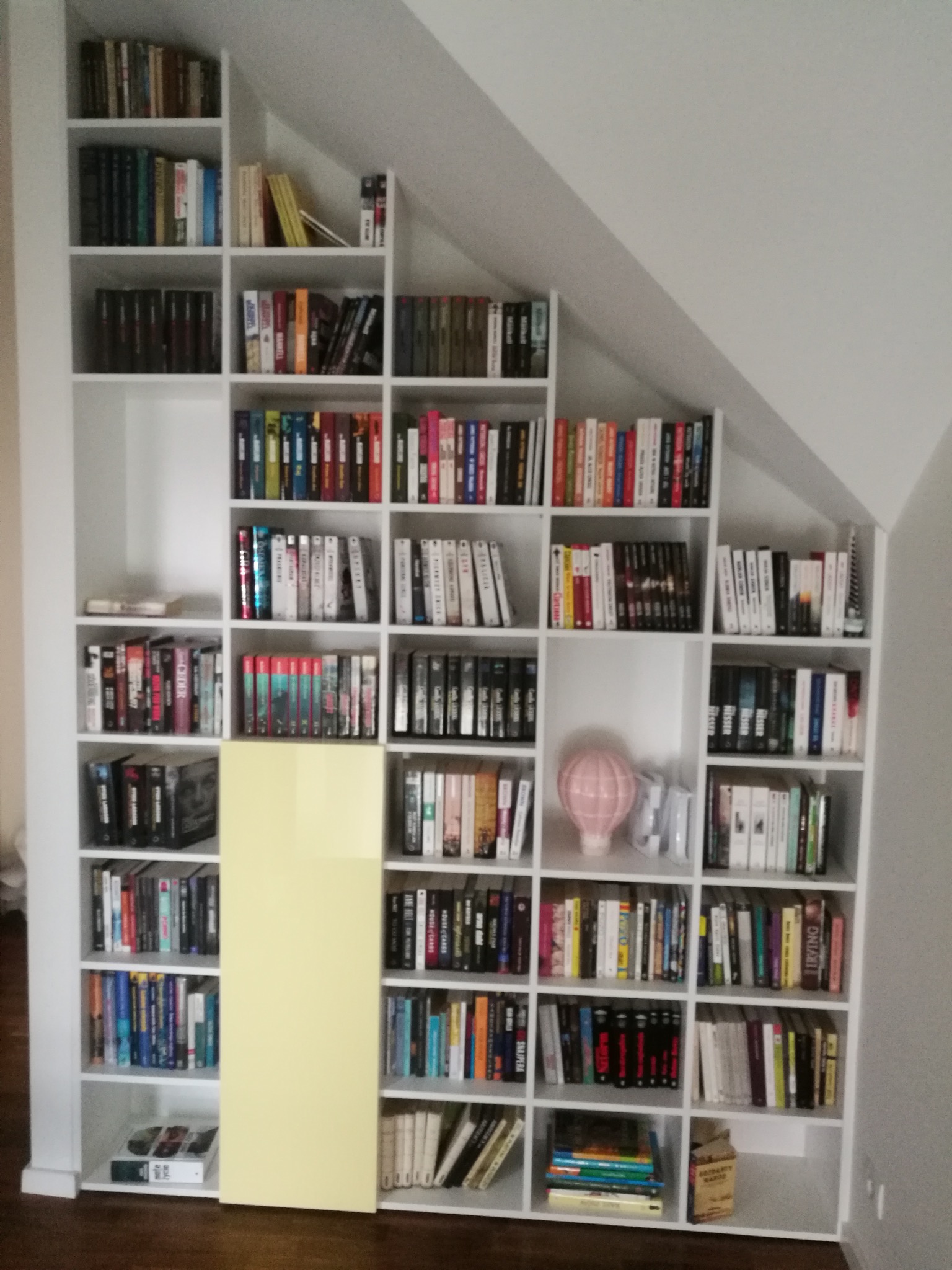 Biały regał na książki, dopasowany do skosu dachu, z żółtym panelem. Półki pełne książek, na jednej dekoracja w kształcie balonu. Widok na wnętrze.