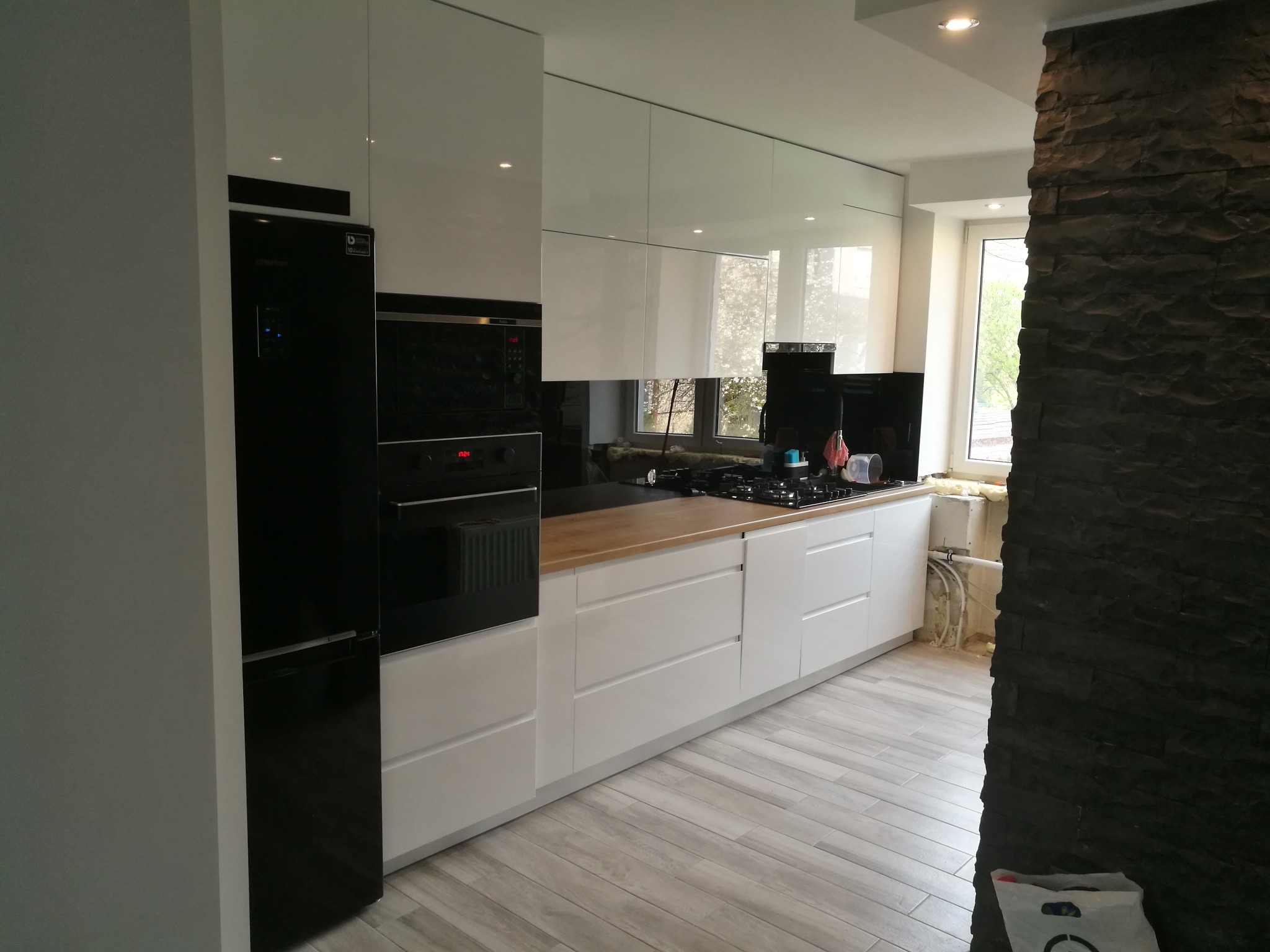 Nowoczesna, biała kuchnia z drewnianym blatem i czarnymi sprzętami AGD. Widoczna ściana z kamienia dekoracyjnego i okno z widokiem na zieleń. Minimalistyczny design.