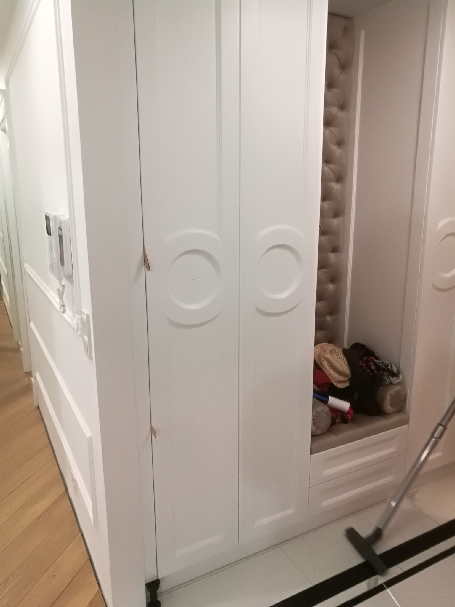 Biała szafa wnękowa z siedziskiem i tapicerowanym panelem w przedpokoju. Widoczne ubrania i akcesoria w otwartej części szafy. Minimalistyczny design.