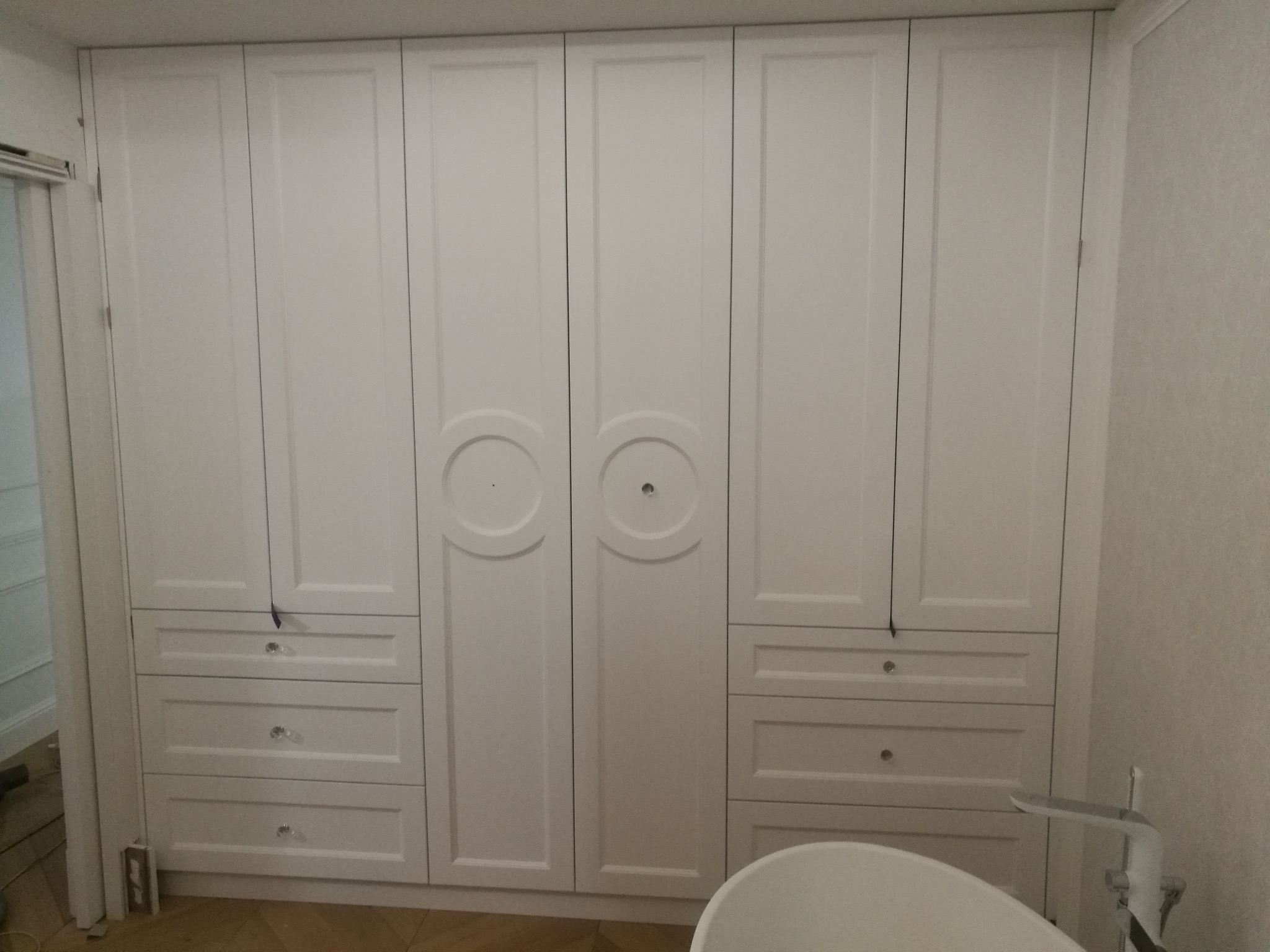 Biała szafa wnękowa z frezowanymi frontami i szufladami z kryształowymi gałkami, widoczna w łazience, z elementami armatury w kadrze.