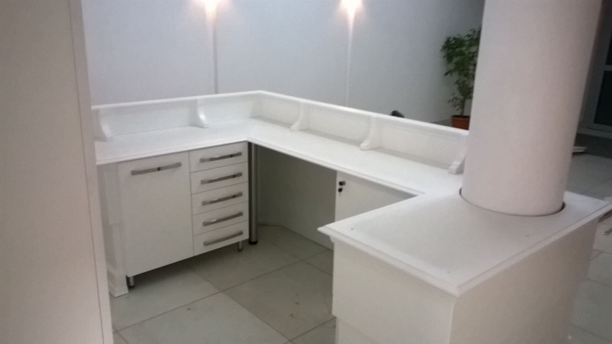 Biała, narożna lada recepcyjna z szafkami i szufladami, oświetlona od góry, z rośliną w tle. Minimalistyczny design, gładkie powierzchnie, widoczny fragment kolumny.