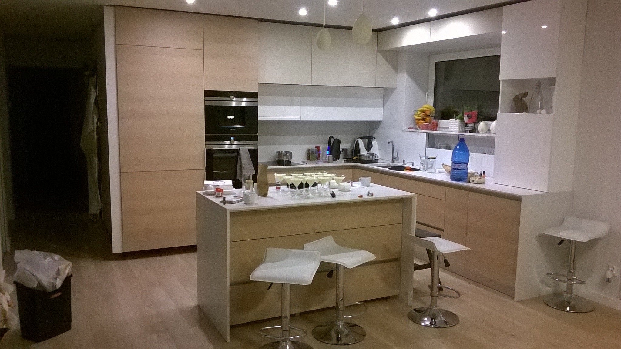 Jasna kuchnia z wyspą i białymi hokerami. Ustawione kieliszki sugerują przygotowania do przyjęcia. Minimalistyczny design, ciepłe oświetlenie.