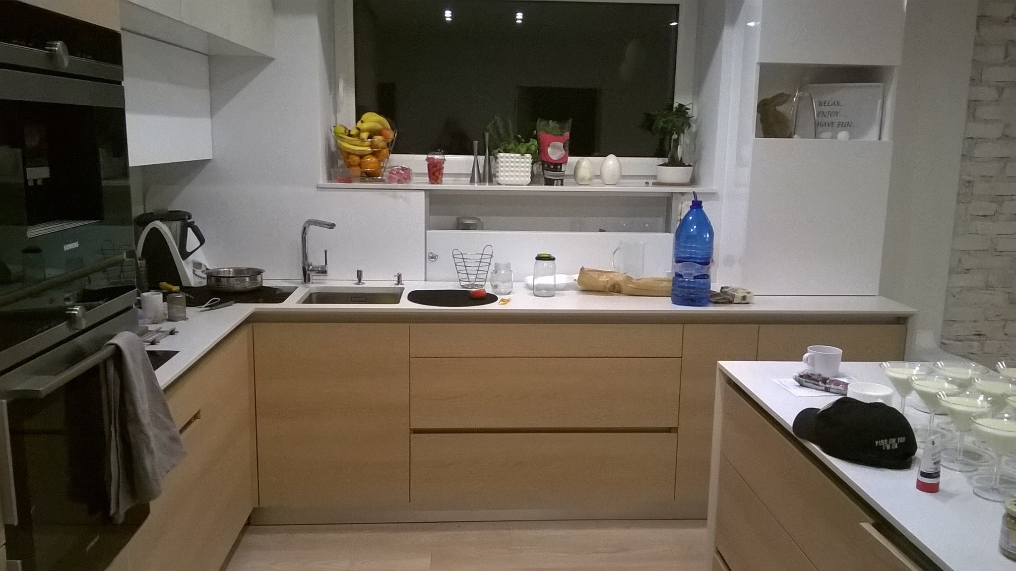 Jasna kuchnia z drewnianymi frontami szafek, widok na okno z dekoracjami. Na blacie naczynia i akcesoria kuchenne, czapka z napisem i szklanki z deserem.