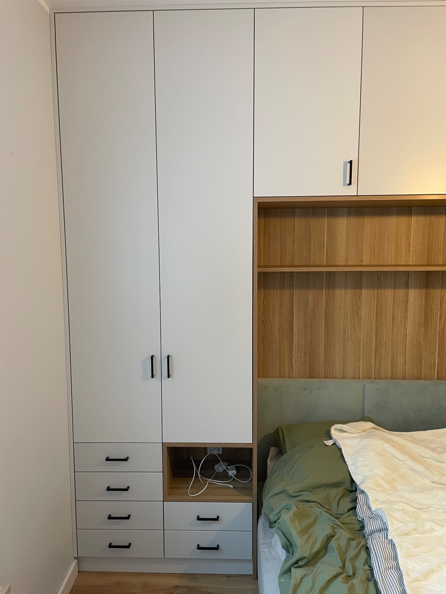 Biała szafa wnękowa z drewnianymi akcentami i szufladami, zintegrowana z wezgłowiem łóżka. Minimalistyczny design, czarne uchwyty, widoczne gniazdko elektryczne w półce.