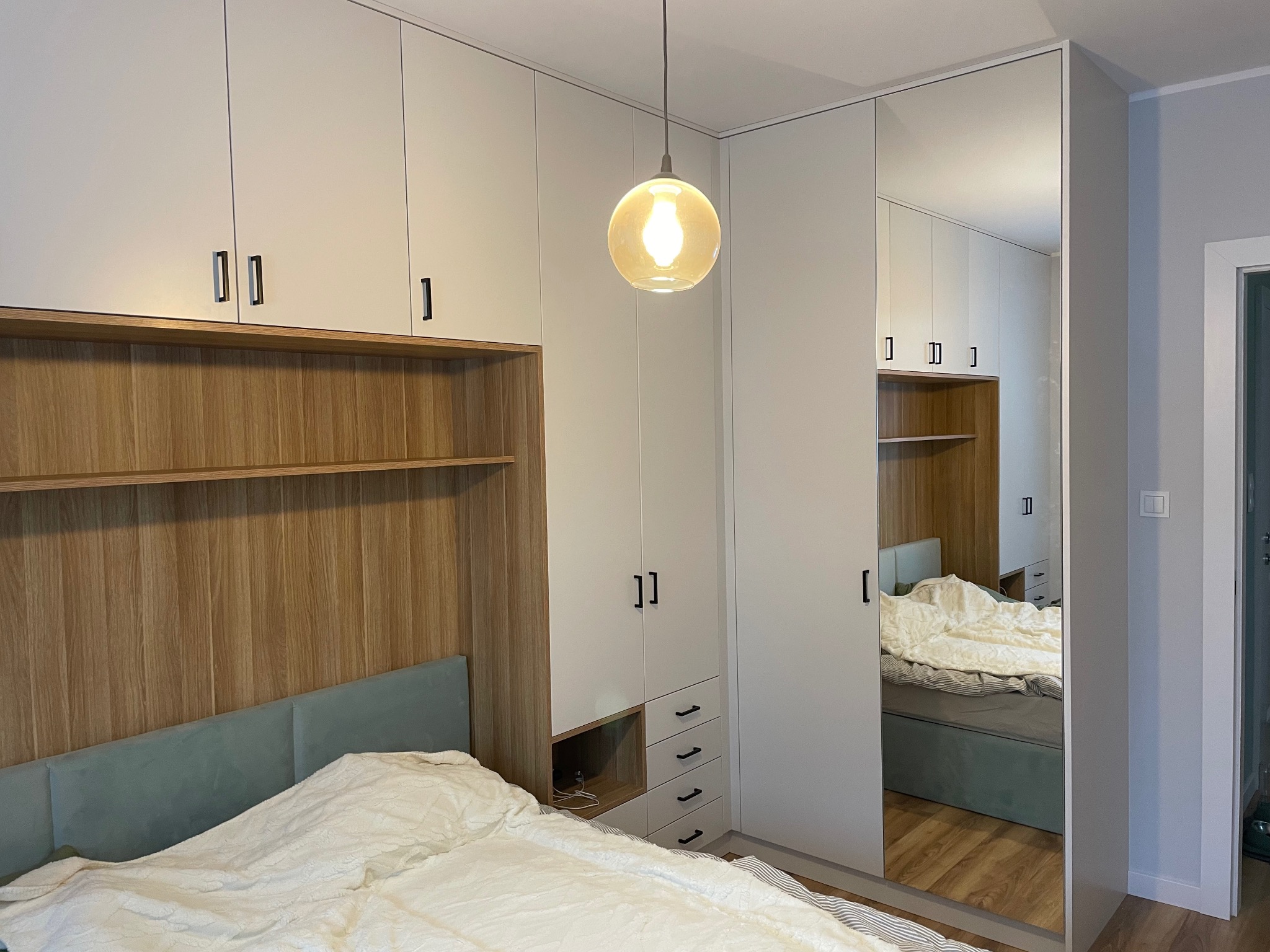 Szafa wnękowa z lustrem i drewnianym wykończeniem nad łóżkiem w sypialni. Minimalistyczny design, wisząca lampa z żarówką Edisona. Widok na odbicie szafy w lustrze.