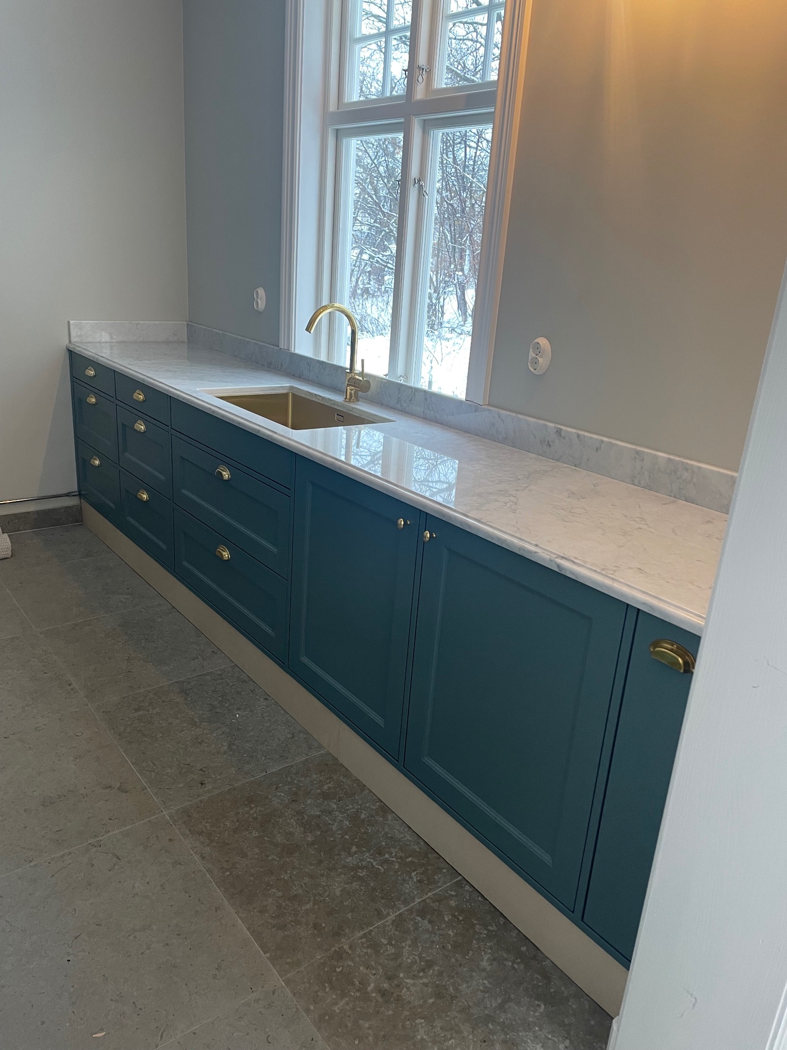 Elegancka zabudowa kuchenna w odcieniach zieleni z marmurowym blatem i złotą armaturą, Wejherowo. Widok na okno z zimowym krajobrazem.