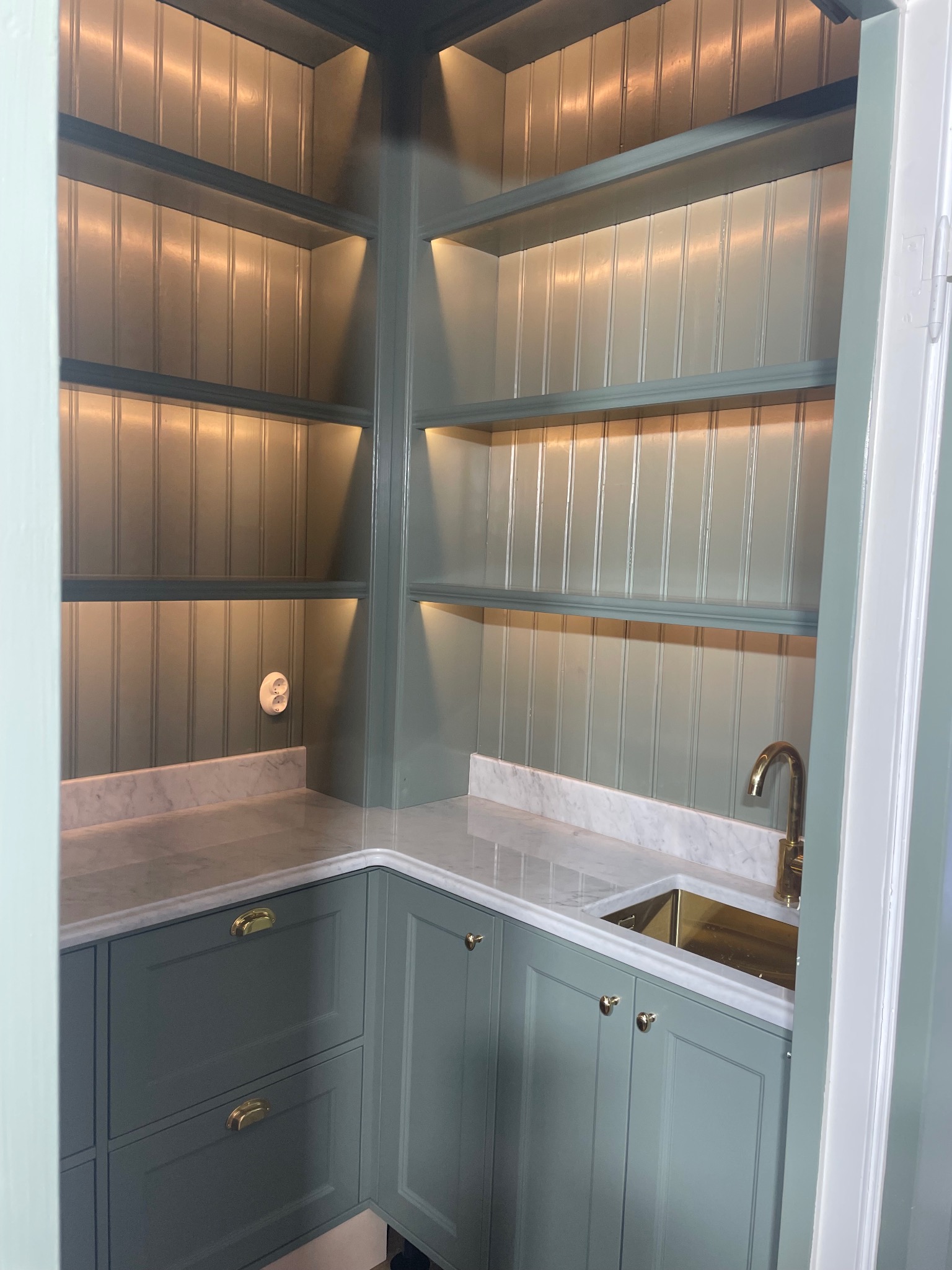 Szara zabudowa kuchenna z marmurowym blatem, złotą armaturą i oświetleniem LED w półkach. Klasyczny styl, widok z perspektywy wejścia do pomieszczenia.