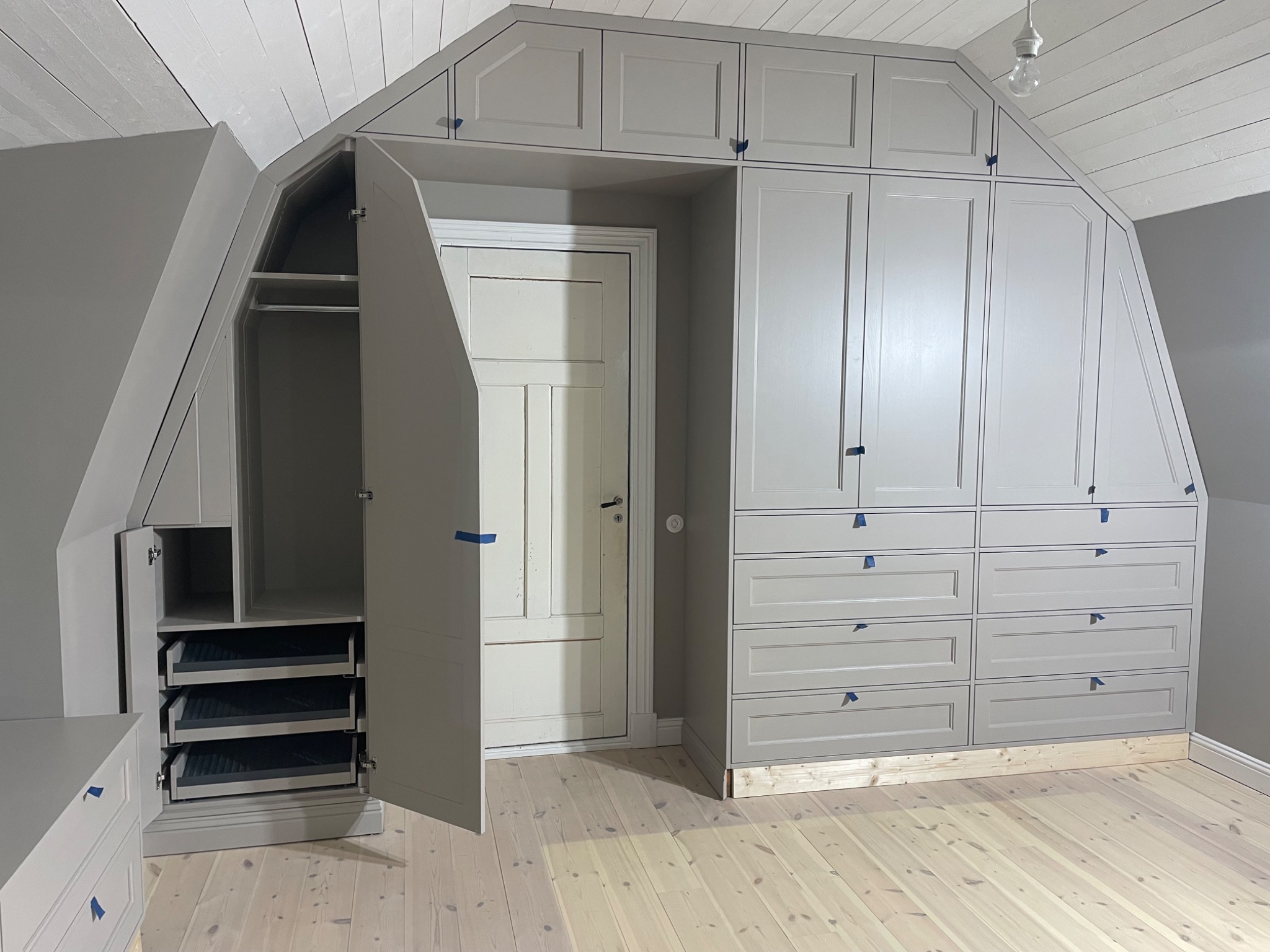 Szara szafa wnękowa na poddaszu z otwartymi drzwiami, ukazująca półki i szuflady. Widoczna drewniana podłoga i białe, skośne panele sufitowe.